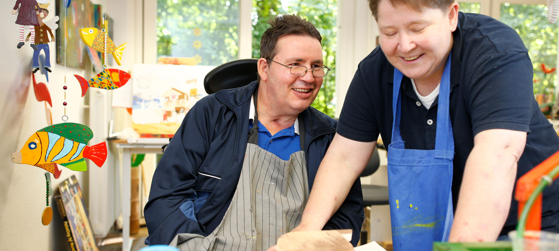Bild zeigt einen Mann mit einer Behinderung und eine Frau, die beide eine Schürze tragen und etwas basteln