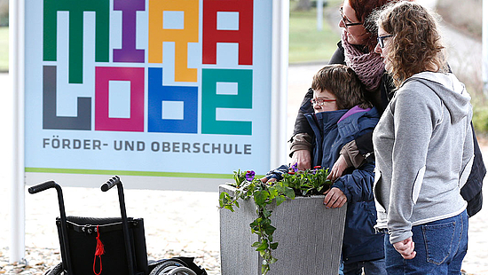 Zwei Frauen und ein Kind stehen vor dem Schild der Mira Lobe Schule. Vorne im Bild ist ein Rollstuhl zu sehen.