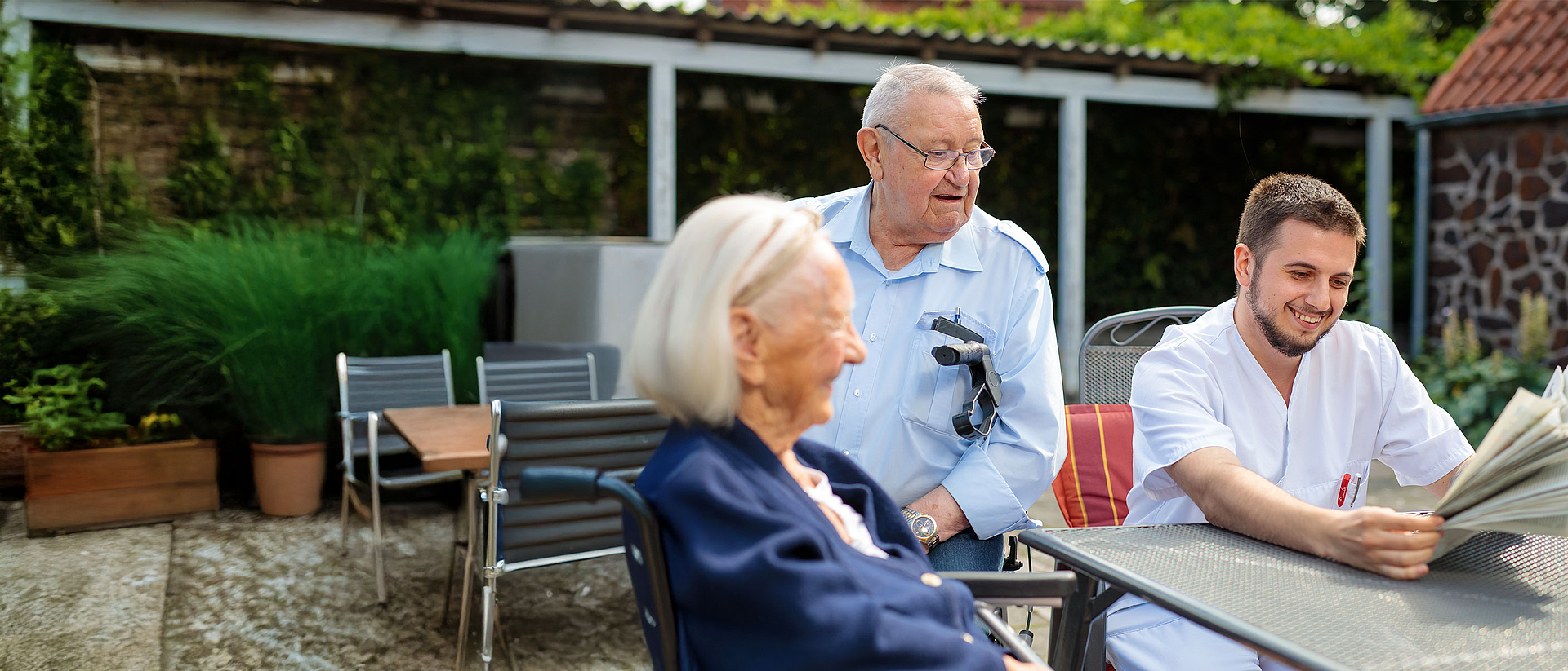 Mitarbeiter der Altenhilfe im Altenzentrum Kirchrode liest zwei Senior*innen im Garten aus der Zeitung vor.