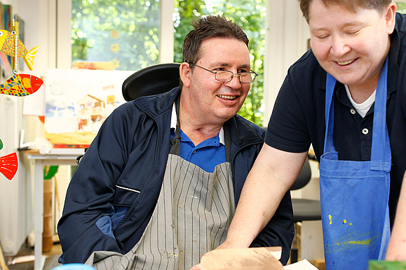 Bild zeigt einen Mann mit einer Behinderung und eine Frau, die beide eine Schürze tragen und etwas basteln